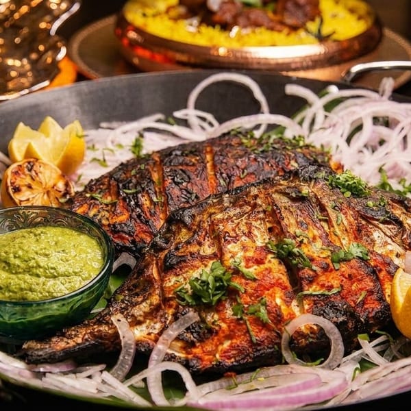 Two whole grilled fish, garnished with onions, lemon, and green chutney, in a black pan.