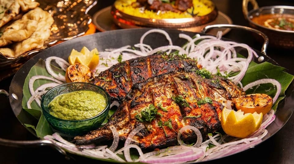 Two whole grilled fish, garnished with onions, lemon, and green chutney, in a black pan.