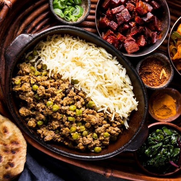 A vibrant Indian thali with keema matar, rice, naan, and various side dishes on a woven tray.