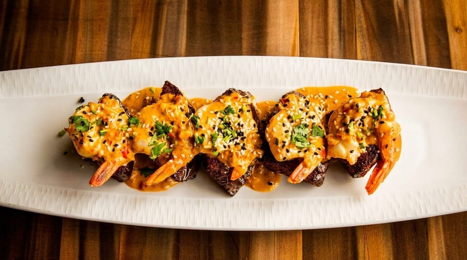 Five pieces of steak topped with shrimp, orange sauce, sesame seeds, and cilantro on a white plate.