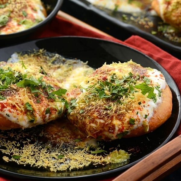 Two plates of Indian chaat, topped with yogurt, spices, and fresh herbs, on a wooden tray.