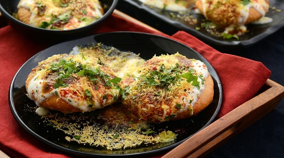Two plates of Indian chaat, topped with yogurt, spices, and fresh herbs, on a wooden tray.